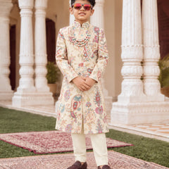 A young boy modeling a traditional Indian outfit consisting of a cream-colored sherwani with colorful paisley patterns, cream trousers, a beaded necklace, and sunglasses, standing in front of a grand building with white pillars.