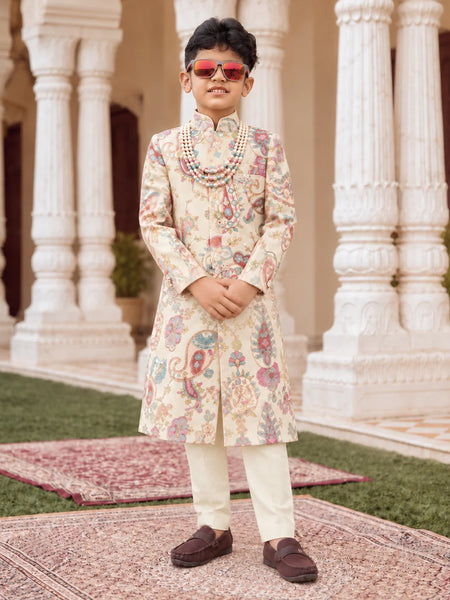 A young boy modeling a traditional Indian outfit consisting of a cream-colored sherwani with colorful paisley patterns, cream trousers, a beaded necklace, and sunglasses, standing in front of a grand building with white pillars.