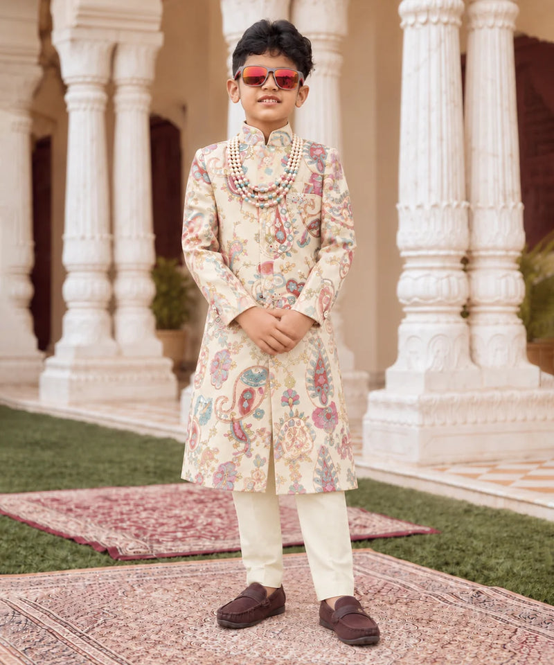 A young boy modeling a traditional Indian outfit consisting of a cream-colored sherwani with colorful paisley patterns, cream trousers, a beaded necklace, and sunglasses, standing in front of a grand building with white pillars.