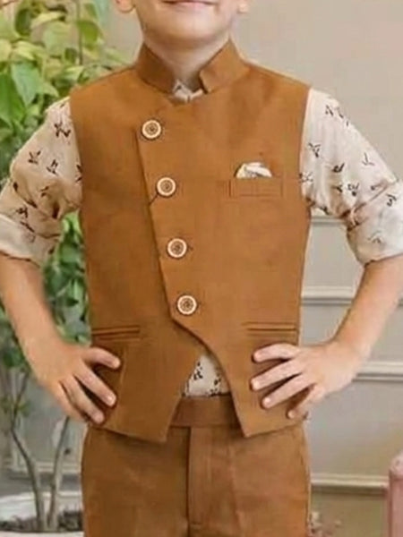 A child wearing a brown Nehru jacket-style vest with four decorative buttons, a chest pocket with a pocket square, and matching brown trousers over a light patterned short-sleeve shirt, standing indoors with hands on hips.