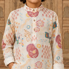 A child wearing a long-sleeve kurta with a white base and colorful traditional Indian print motifs, set against a warm, ornate architectural background.