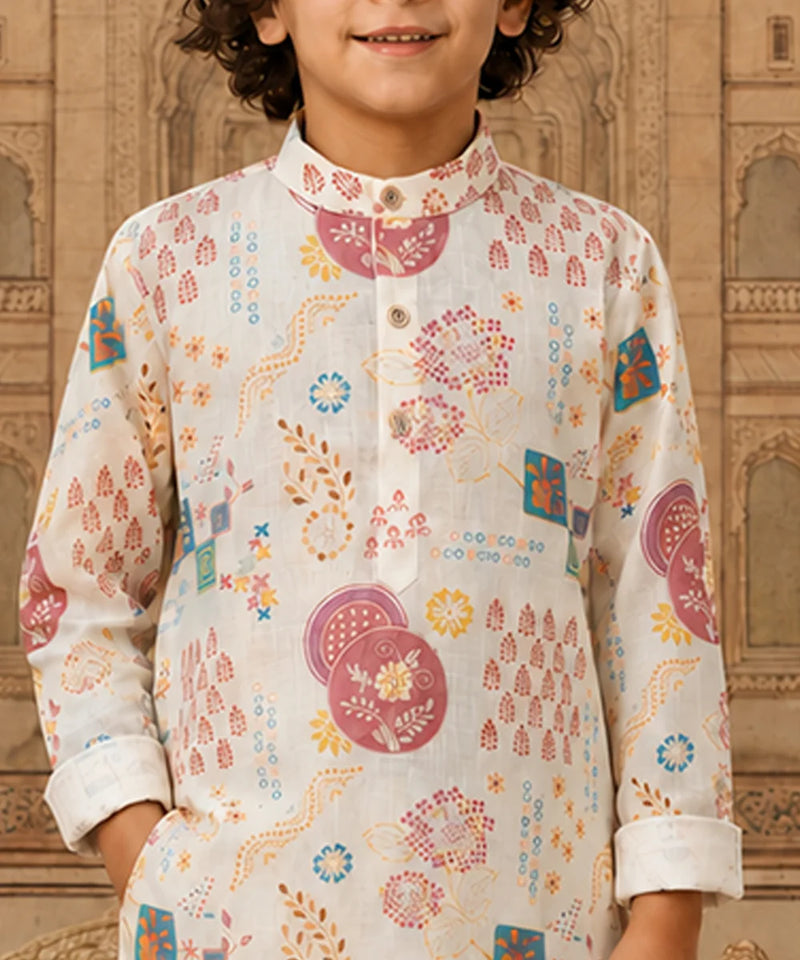 A child wearing a long-sleeve kurta with a white base and colorful traditional Indian print motifs, set against a warm, ornate architectural background.