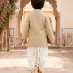 A toddler viewed from behind wearing a beige kurta and white dhoti pants, walking in a courtyard with arches and flowers in the background.