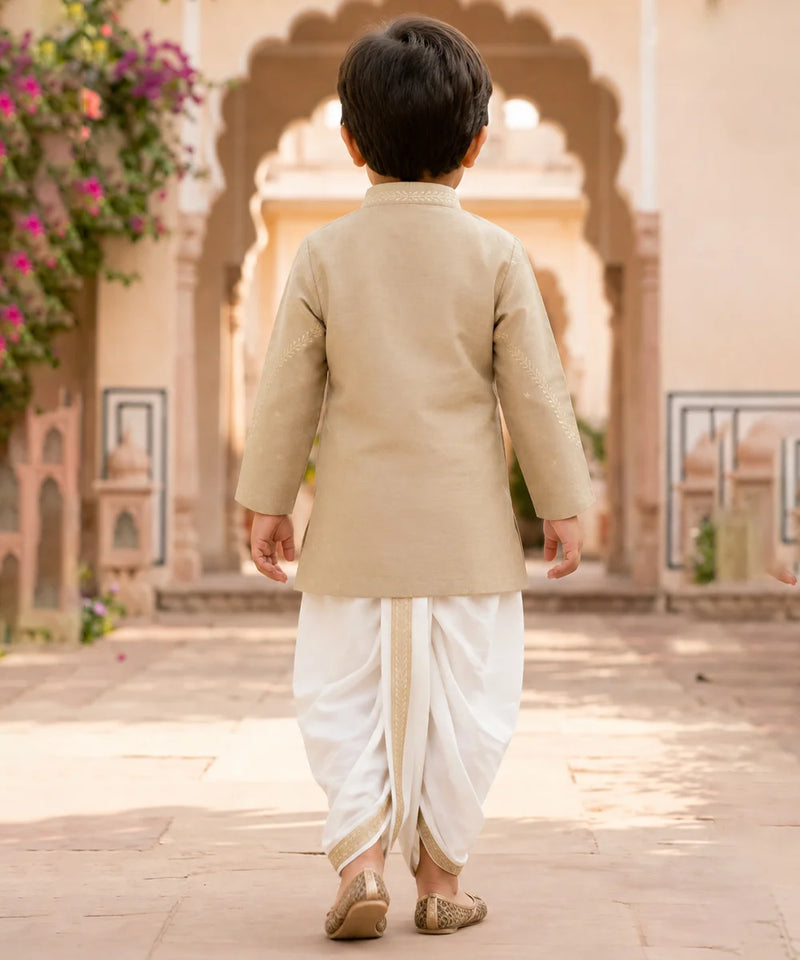 A toddler viewed from behind wearing a beige kurta and white dhoti pants, walking in a courtyard with arches and flowers in the background.