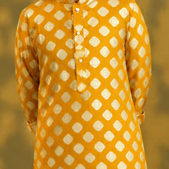 A child viewed from behind wearing a mustard yellow kurta with a white geometric diamond pattern, paired with white trousers and sandals, standing against a warm yellow background with soft shadow details.