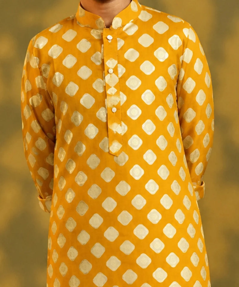 A child viewed from behind wearing a mustard yellow kurta with a white geometric diamond pattern, paired with white trousers and sandals, standing against a warm yellow background with soft shadow details.