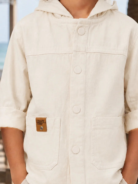 A close-up of a person wearing a cream-colored hooded button-up shirt with front pockets and rolled-up sleeves, photographed outdoors near a blurred beach or railing background.