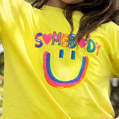 A close-up of a person wearing a bright yellow t-shirt with a colorful graphic that reads 'SOMEBODY' in rainbow letters and a matching smiley face underneath.