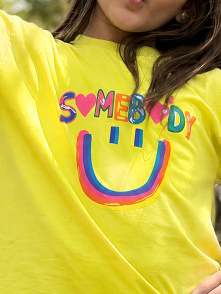 A close-up of a person wearing a bright yellow t-shirt with a colorful graphic that reads 'SOMEBODY' in rainbow letters and a matching smiley face underneath.