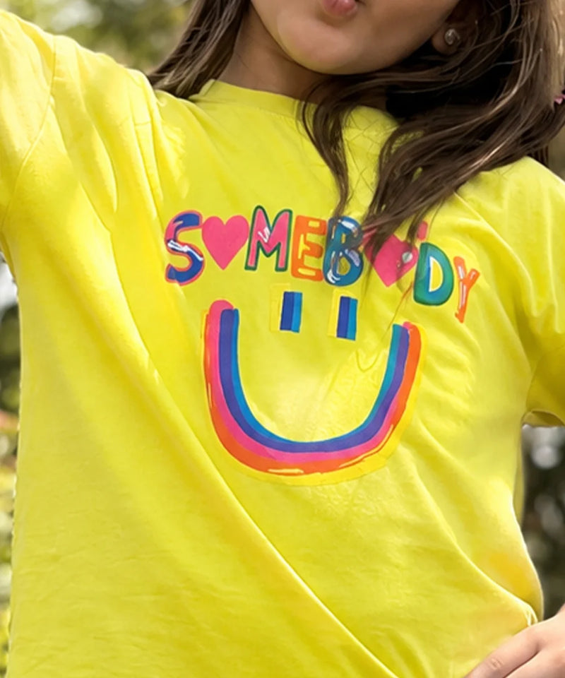A close-up of a person wearing a bright yellow t-shirt with a colorful graphic that reads 'SOMEBODY' in rainbow letters and a matching smiley face underneath.