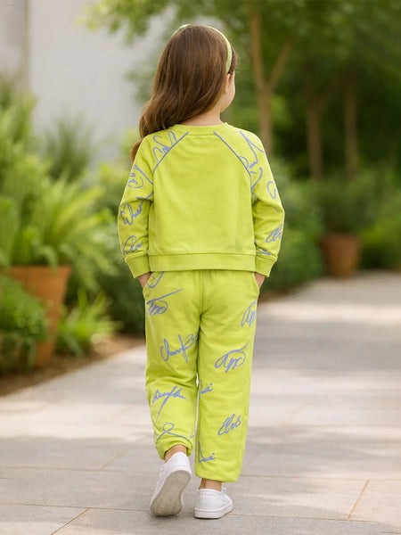 Child wearing a bright green outfit with blue patterns, standing on a pathway with plants in the background.