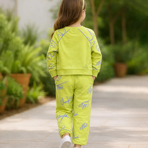 Child wearing a bright green outfit with blue patterns, standing on a pathway with plants in the background.