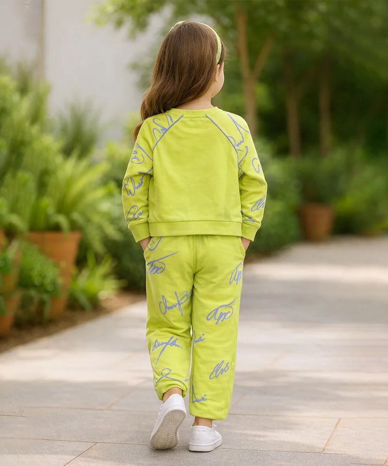 Child wearing a bright green outfit with blue patterns, standing on a pathway with plants in the background.