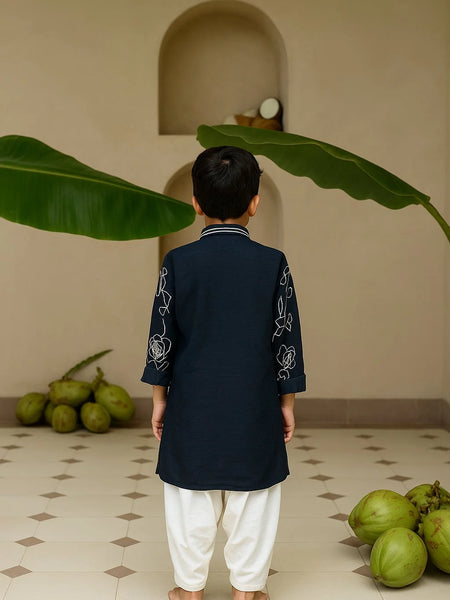 Child wearing a dark blue kurta with white embroidery in a room with green fruits and leaves.