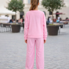 Person wearing a pink tracksuit standing on a paved area with trees and people in the background.