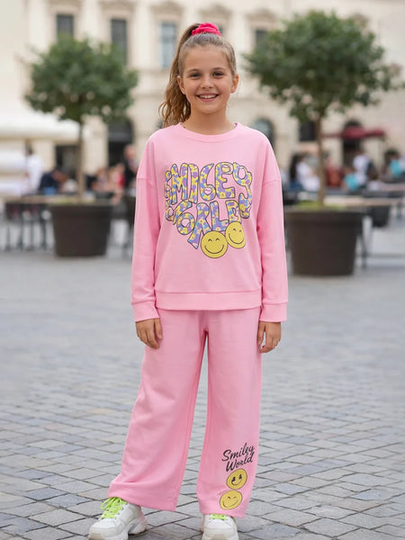 Young girl wearing a pink 'Smiley World' outfit standing on a paved area with trees and people in the background.