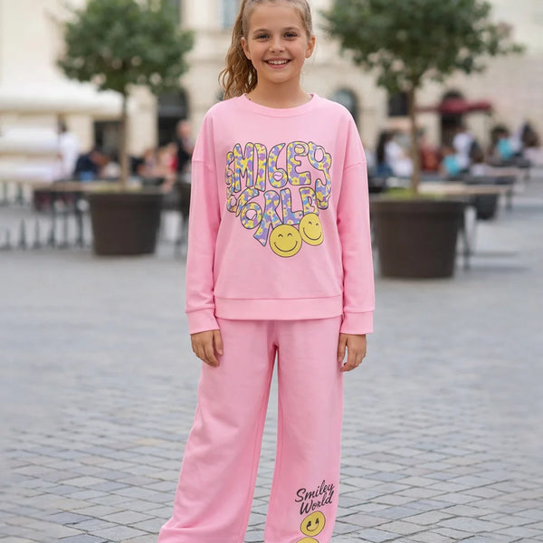 Young girl wearing a pink 'Smiley World' outfit standing on a paved area with trees and people in the background.