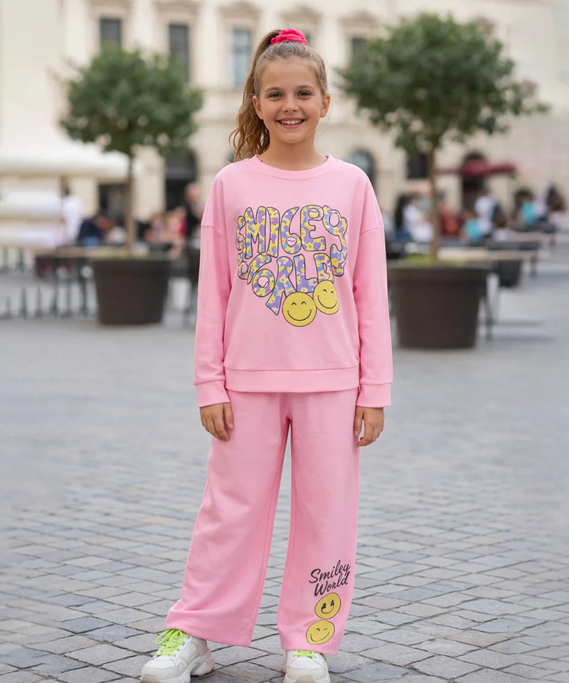 Young girl wearing a pink 'Smiley World' outfit standing on a paved area with trees and people in the background.