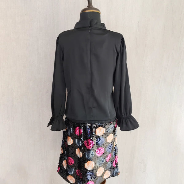 Black blouse with ruffled sleeves on a mannequin wearing a colorful skirt against a plain background