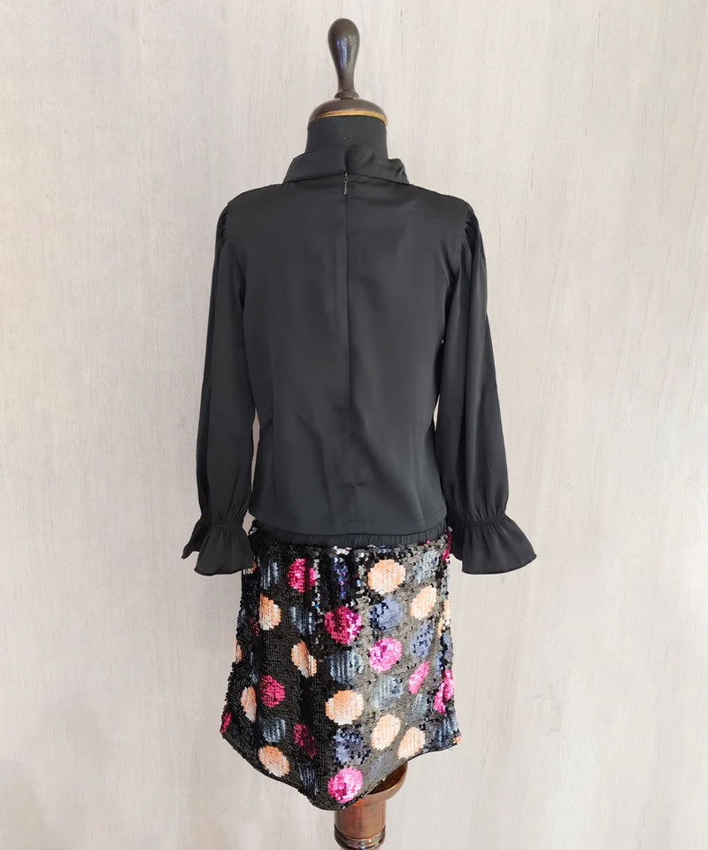 Black blouse with ruffled sleeves on a mannequin wearing a colorful skirt against a plain background