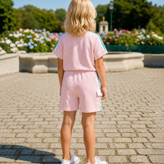 A young girl with blonde hair is seen from behind, wearing a light pink two-piece tracksuit with light blue stripes on the sleeves and shorts, paired with white sneakers, standing on a stone paved path in a park.