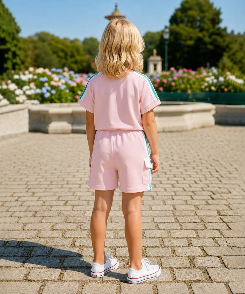 A young girl with blonde hair is seen from behind, wearing a light pink two-piece tracksuit with light blue stripes on the sleeves and shorts, paired with white sneakers, standing on a stone paved path in a park.