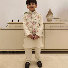 A boy wearing a cream colored kurta set with a printed waist coat standing in front of a white background.