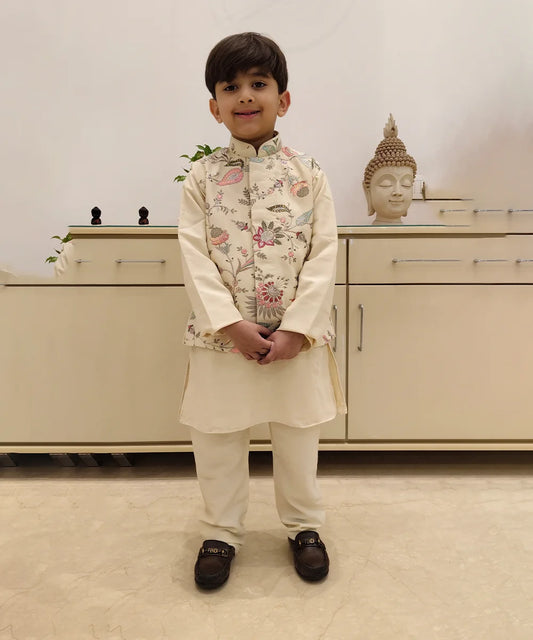 A boy wearing a cream colored kurta set with a printed waist coat standing in front of a white background.