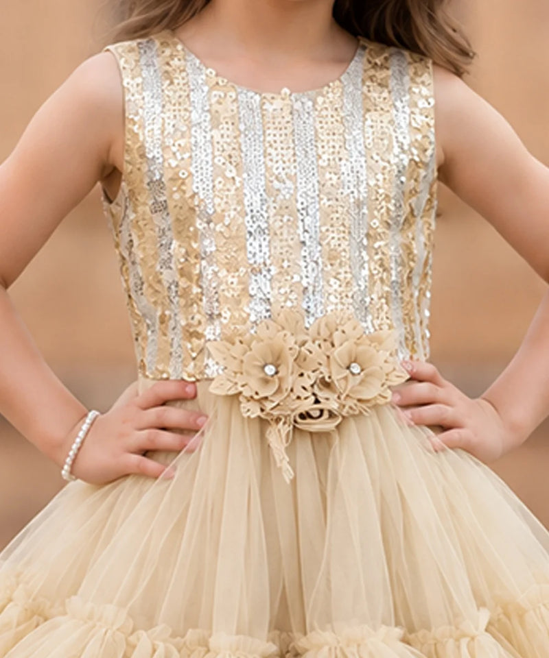 A sleeveless champagne beige and silver sequined dress with a floral waistband and tulle skirt, shown on a model against a warm beige background.