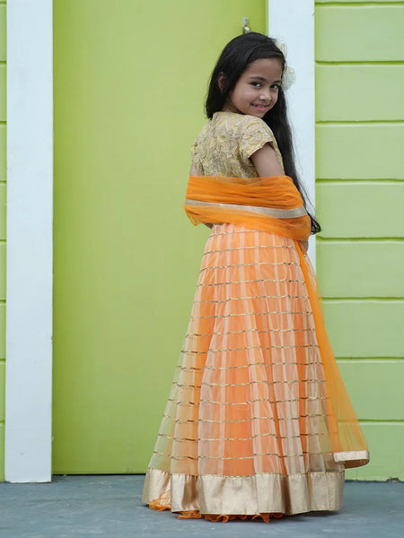 Young girl in an orange and gold traditional outfit standing against a green wall.