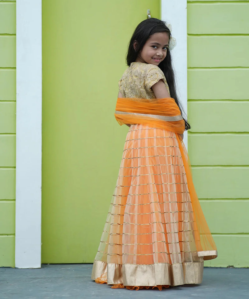 Young girl in an orange and gold traditional outfit standing against a green wall.