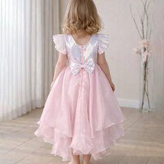 Young girl wearing a pink dress with a white sequin bow back, standing in a room with light-colored walls and wooden floor.