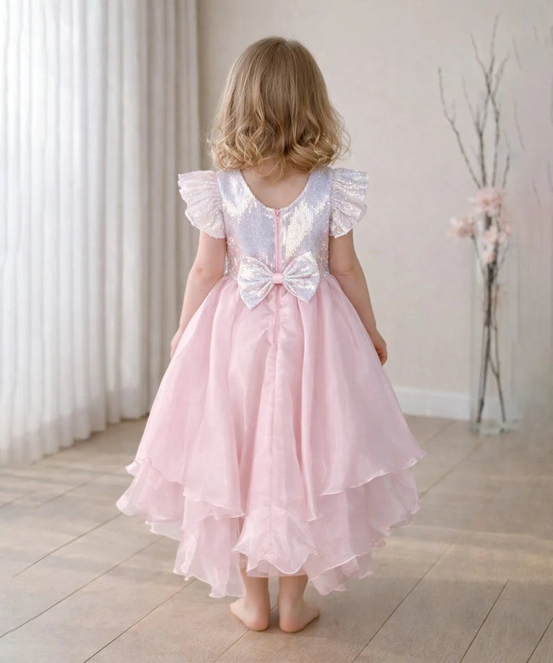 Young girl wearing a pink dress with a white sequin bow back, standing in a room with light-colored walls and wooden floor.