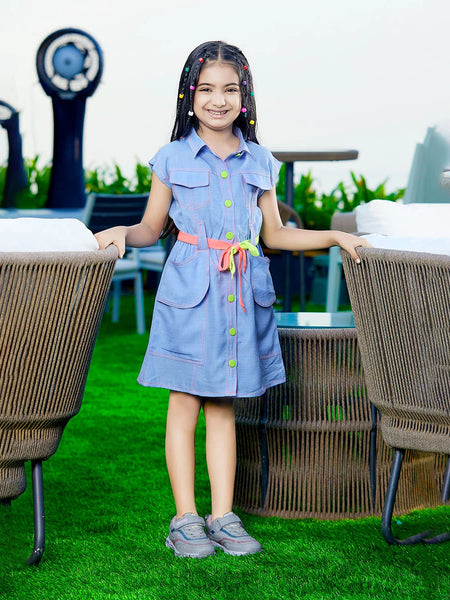 A girl wearing a blue denim party wear dress with long sleeves, standing on a grass-like surface with two large decorative seashells in the background.