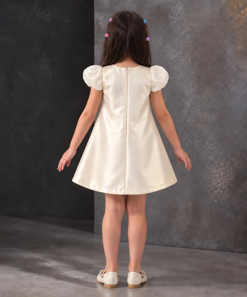 A young girl viewed from behind wearing a cream-colored satin dress with puffed short sleeves and a back zipper, standing against a textured dark gray studio backdrop.