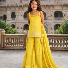 A young girl stands in a courtyard wearing a bright yellow traditional outfit with a sleeveless top, wide-leg pants, and a matching draped scarf. The outfit has white embroidered details and is photographed against a large stone building in the background.
