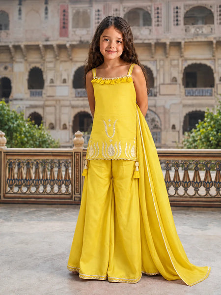 A young girl stands in a courtyard wearing a bright yellow traditional outfit with a sleeveless top, wide-leg pants, and a matching draped scarf. The outfit has white embroidered details and is photographed against a large stone building in the background.