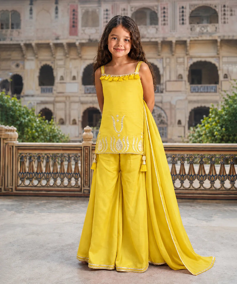 A young girl stands in a courtyard wearing a bright yellow traditional outfit with a sleeveless top, wide-leg pants, and a matching draped scarf. The outfit has white embroidered details and is photographed against a large stone building in the background.