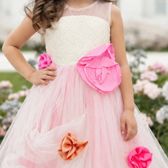 A young girl wearing a sleeveless pink and white tulle flower girl dress with large pink and orange fabric flowers, photographed outdoors in a garden setting.