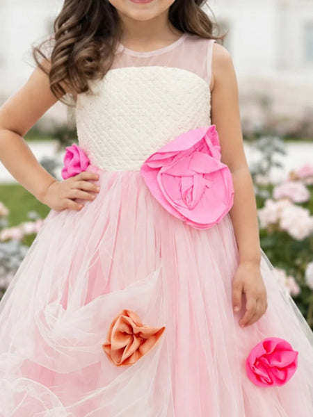 A young girl wearing a sleeveless pink and white tulle flower girl dress with large pink and orange fabric flowers, photographed outdoors in a garden setting.