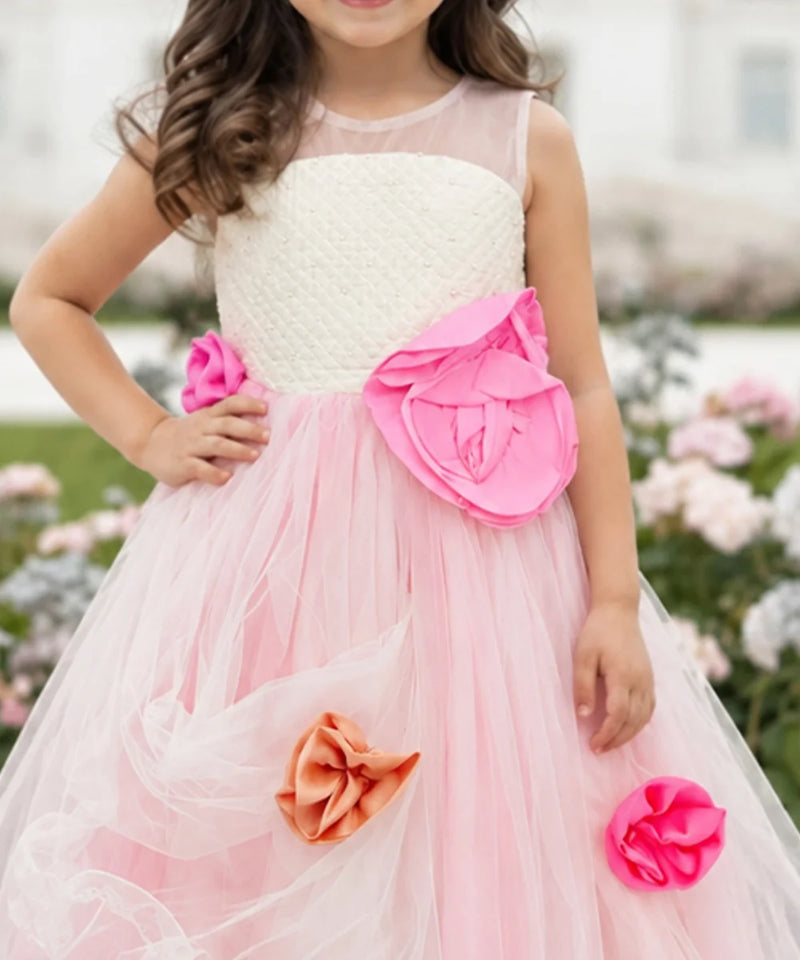 A young girl wearing a sleeveless pink and white tulle flower girl dress with large pink and orange fabric flowers, photographed outdoors in a garden setting.
