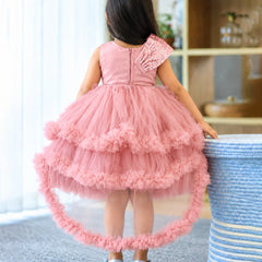 A young girl viewed from behind wearing a pink high-low tulle dress with a ruffled asymmetrical shoulder and a flared tiered skirt, standing indoors next to a blue-and-white woven stool.