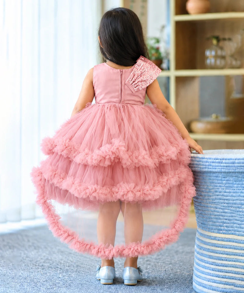 A young girl viewed from behind wearing a pink high-low tulle dress with a ruffled asymmetrical shoulder and a flared tiered skirt, standing indoors next to a blue-and-white woven stool.