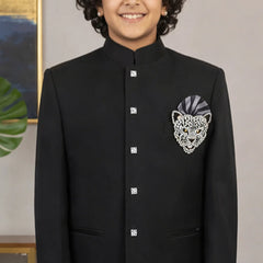 Young boy wearing a black traditional outfit with a decorative patch, standing against a neutral background.