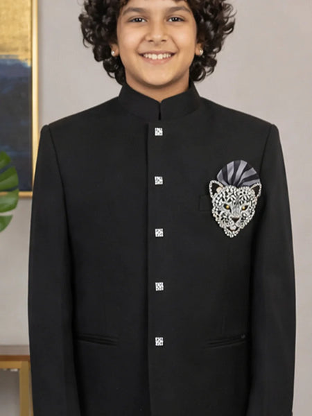Young boy wearing a black traditional outfit with a decorative patch, standing against a neutral background.