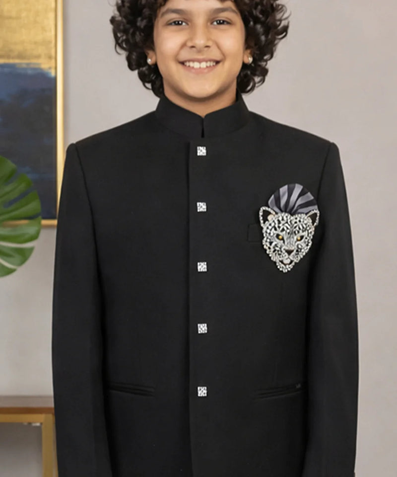 Young boy wearing a black traditional outfit with a decorative patch, standing against a neutral background.