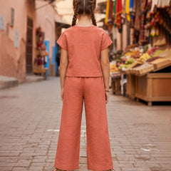 A child standing on a cobblestone street, viewed from behind, wearing a matching terracotta-colored short-sleeve top and wide-leg pants set. The background shows a blurred outdoor market scene with stalls and colorful flags.