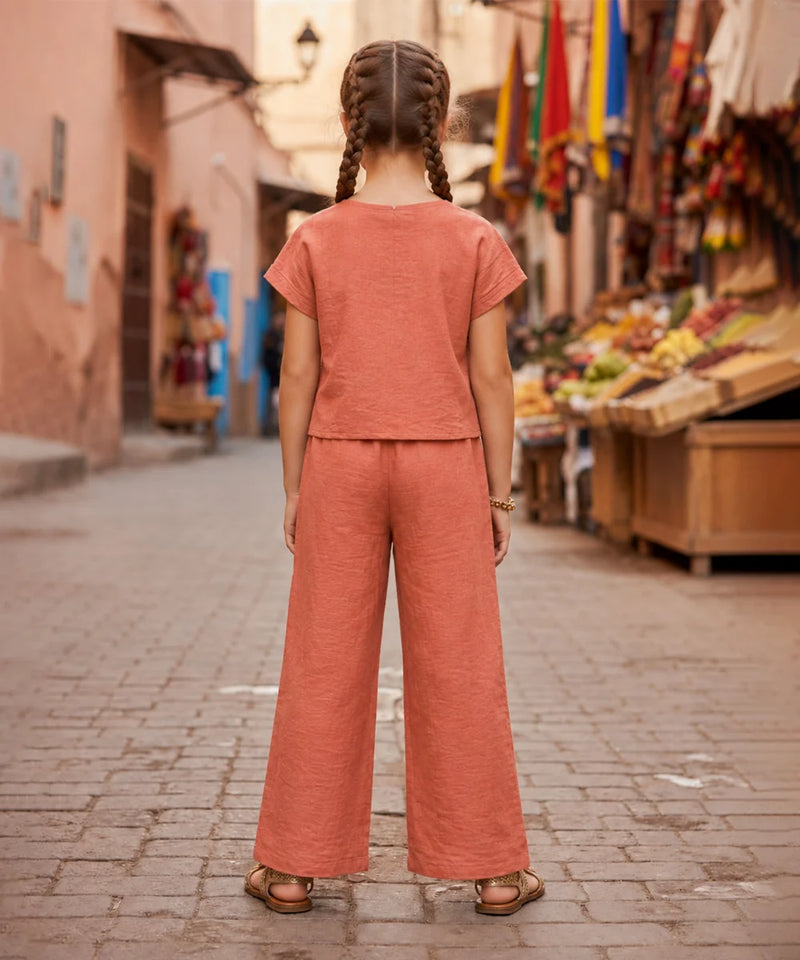 A child standing on a cobblestone street, viewed from behind, wearing a matching terracotta-colored short-sleeve top and wide-leg pants set. The background shows a blurred outdoor market scene with stalls and colorful flags.