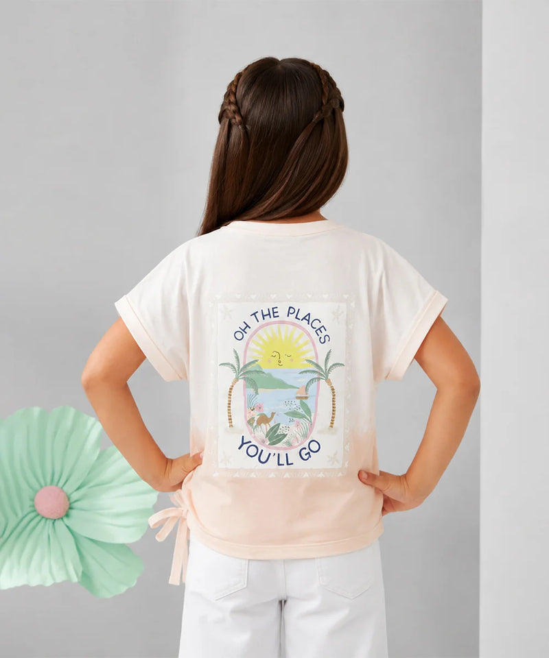 A young girl is shown from behind wearing a cream-colored t-shirt with a colorful tropical beach graphic and the text 'OH THE PLACE'S YOU'LL GO'. She is paired with white pants against a neutral gray background with a decorative mint green paper flower on the left.