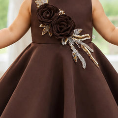 A sleeveless brown dress with a large dark brown fabric flower and gold and silver embellished leaves on the bodice, worn by a child.